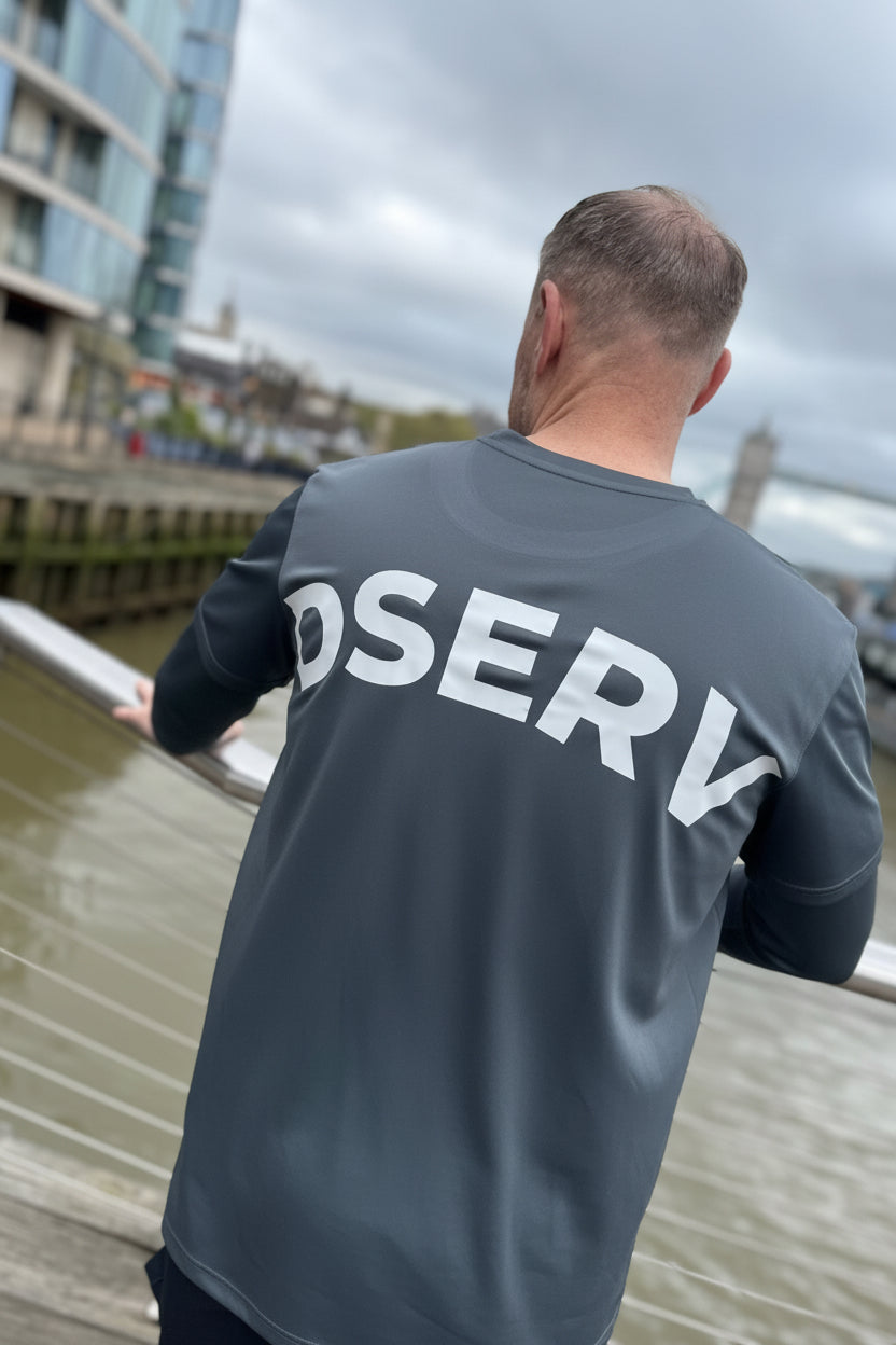 Man wearing a dark gray jacket with 'DSERV' on the back, standing by a waterfront with London buildings in the background.