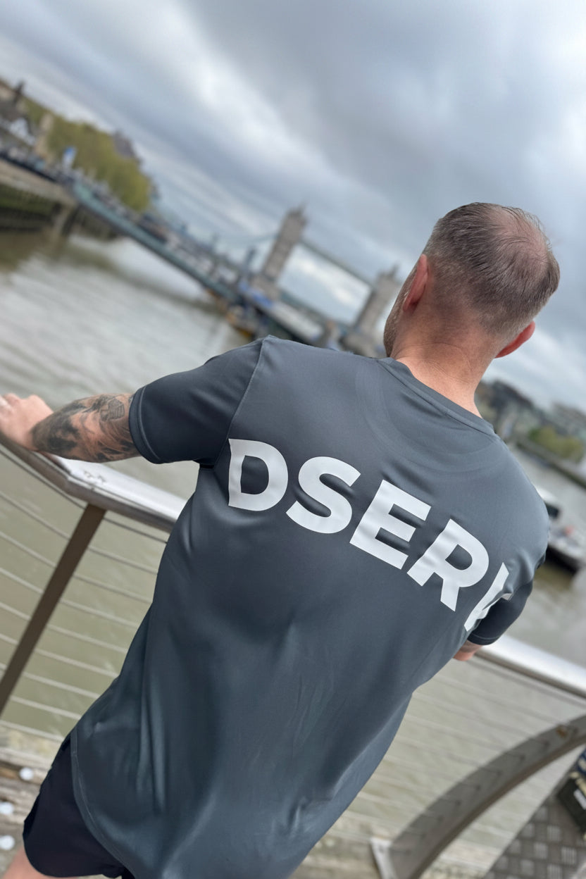 Person wearing a gray t-shirt with 'DSERV' on the back, standing by a waterfront with the Tower Bridge in the back ground