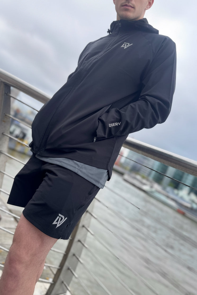 Person wearing a black jacket and shorts with a logo, standing on a bridge.