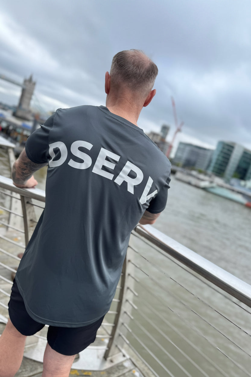 Person wearing a gray t-shirt with 'DSERV' on the back, standing on a bridge overlooking a cityscape.