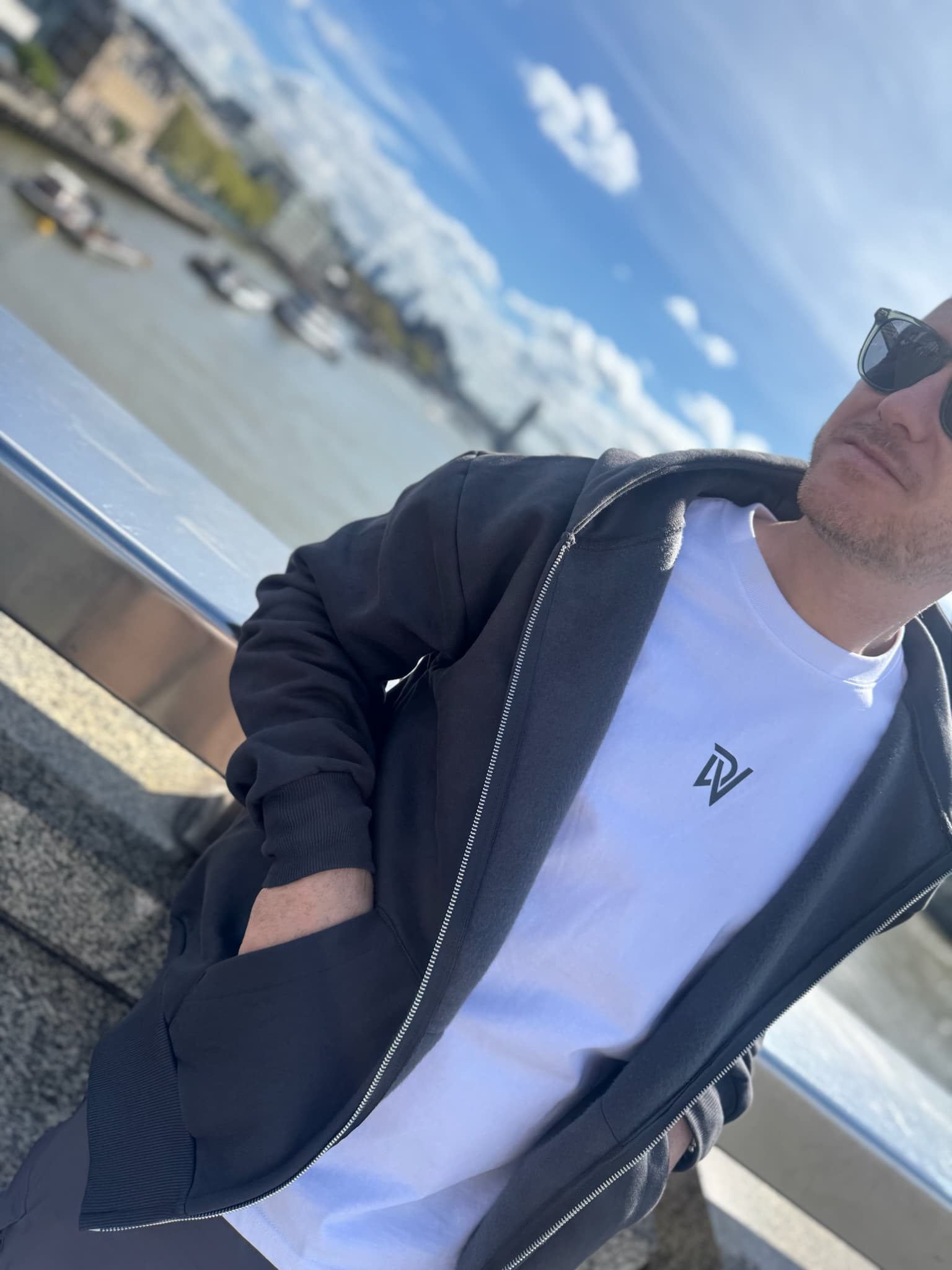 Man wearing sunglasses and a dark jacket with a white shirt featuring a logo, standing by a waterfront.
