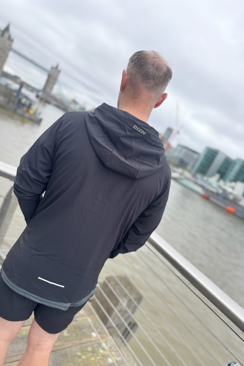 Person wearing a black jacket with a brand logo, standing by a waterfront with cityscape in the background.