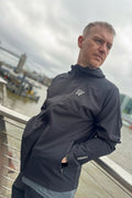 Man wearing a black jacket with a DV logo, standing on a bridge with a cityscape background.